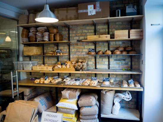 Shelves are stocked with various types of bread and pastries, displayed in a rustic setting with a brick wall backdrop. There are loaves of baked bread, croissants, and other bakery items on wooden shelves. Bags of flour and supplies are stacked below the shelves. A metal light fixture hangs from the ceiling, illuminating the products.