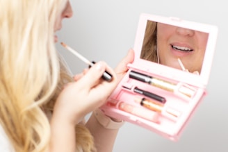 A person is applying lip gloss using a small makeup kit with a built-in mirror. The kit contains various cosmetics, including lip gloss and possibly mascara or eyeliner tubes. The reflection shows the person's lips as they apply the product. The scene is close-up and focused on the action of applying makeup.