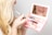 A person is applying lip gloss using a small makeup kit with a built-in mirror. The kit contains various cosmetics, including lip gloss and possibly mascara or eyeliner tubes. The reflection shows the person's lips as they apply the product. The scene is close-up and focused on the action of applying makeup.