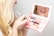 A person is applying lip gloss using a small makeup kit with a built-in mirror. The kit contains various cosmetics, including lip gloss and possibly mascara or eyeliner tubes. The reflection shows the person's lips as they apply the product. The scene is close-up and focused on the action of applying makeup.
