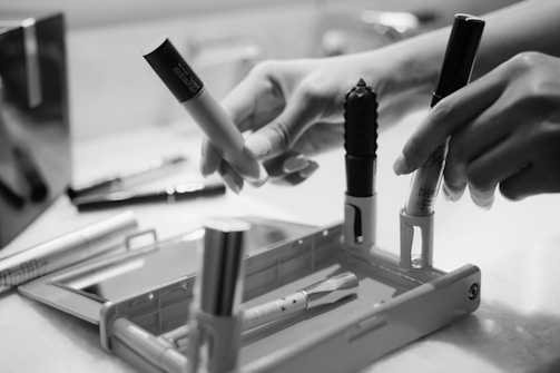 Hands are interacting with several makeup items, including tubes of lipstick and mascara, placed on a rectangular tray with a mirror. The image presents a scene of personal grooming or beauty routine in a bathroom setting.
