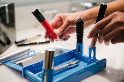 Hands holding various makeup products, including a red and a pink lipstick, over a blue makeup organizer. The background features a mirror and a blurred out countertop with more beauty items.