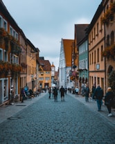 A picturesque European cobblestone street stretches through a charming town, flanked by colorful buildings with flower-adorned facades. People leisurely walk along the street, adding a lively atmosphere. The architecture is a mix of traditional half-timbered and colorful plaster structures, with iron signs hanging outside various businesses.