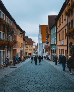 A picturesque European cobblestone street stretches through a charming town, flanked by colorful buildings with flower-adorned facades. People leisurely walk along the street, adding a lively atmosphere. The architecture is a mix of traditional half-timbered and colorful plaster structures, with iron signs hanging outside various businesses.
