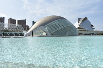 Futuristic architecture is featured prominently, with sleek, curved structures surrounded by a reflective body of water. The buildings have modern, glass-like exteriors. In the background, there are several high-rise buildings with a more traditional facade.