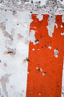 A textured wall with chipped paint showcasing an aged surface. The colors include weathered white and bright orange, with visible cracks and peeling revealing the rough texture beneath.