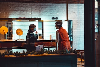 Two people are interacting inside a store, with one seated behind a counter wearing a mask and the other standing opposite wearing a face shield. The store features a brick wall painted white and illuminated glass cases. The atmosphere is warm and inviting, with decorative elements hinting at a festive setting.