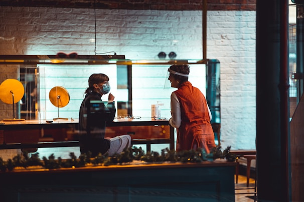 Two people are interacting inside a store, with one seated behind a counter wearing a mask and the other standing opposite wearing a face shield. The store features a brick wall painted white and illuminated glass cases. The atmosphere is warm and inviting, with decorative elements hinting at a festive setting.