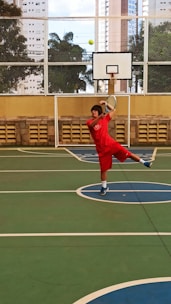 A person in red sportswear is playing tennis on an outdoor court surrounded by high-rise buildings. They are captured mid-action, swinging a racket to hit a green tennis ball. The setting includes a basketball hoop and a wire fence with leafy trees visible in the background.