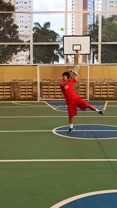 A person in red sportswear is playing tennis on an outdoor court surrounded by high-rise buildings. They are captured mid-action, swinging a racket to hit a green tennis ball. The setting includes a basketball hoop and a wire fence with leafy trees visible in the background.
