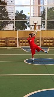 A person in red sportswear is playing tennis on an outdoor court surrounded by high-rise buildings. They are captured mid-action, swinging a racket to hit a green tennis ball. The setting includes a basketball hoop and a wire fence with leafy trees visible in the background.