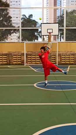 A person in red sportswear is playing tennis on an outdoor court surrounded by high-rise buildings. They are captured mid-action, swinging a racket to hit a green tennis ball. The setting includes a basketball hoop and a wire fence with leafy trees visible in the background.