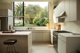 A modern kitchen interior featuring sleek cabinetry and a bright, spacious countertop layout. A large window reveals lush greenery outside, enhancing the natural light flooding the room. Appliances like a stovetop and an oven are integrated seamlessly into the design. A stool and kitchen accessories like a cutting board and a potted plant decorate the space.