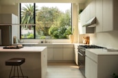 A modern kitchen interior featuring sleek cabinetry and a bright, spacious countertop layout. A large window reveals lush greenery outside, enhancing the natural light flooding the room. Appliances like a stovetop and an oven are integrated seamlessly into the design. A stool and kitchen accessories like a cutting board and a potted plant decorate the space.