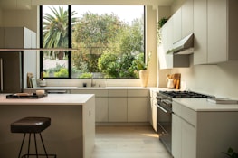 A modern kitchen interior featuring sleek cabinetry and a bright, spacious countertop layout. A large window reveals lush greenery outside, enhancing the natural light flooding the room. Appliances like a stovetop and an oven are integrated seamlessly into the design. A stool and kitchen accessories like a cutting board and a potted plant decorate the space.