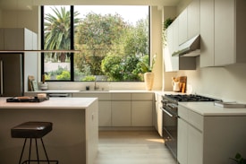 A modern kitchen interior featuring sleek cabinetry and a bright, spacious countertop layout. A large window reveals lush greenery outside, enhancing the natural light flooding the room. Appliances like a stovetop and an oven are integrated seamlessly into the design. A stool and kitchen accessories like a cutting board and a potted plant decorate the space.