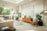 A modern, spacious living room with an open-plan kitchen. It features a light color palette with neutral tones. The kitchen area has sleek, minimalist cabinetry, a stove, and a large window allowing natural light to pour in. The living area includes a comfortable sofa, a circular coffee table, and a wooden sideboard with decorative items. Large leafy plants add a touch of greenery. The room is well-lit with a high ceiling and contemporary decor.