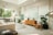 A modern, spacious living room with an open-plan kitchen. It features a light color palette with neutral tones. The kitchen area has sleek, minimalist cabinetry, a stove, and a large window allowing natural light to pour in. The living area includes a comfortable sofa, a circular coffee table, and a wooden sideboard with decorative items. Large leafy plants add a touch of greenery. The room is well-lit with a high ceiling and contemporary decor.