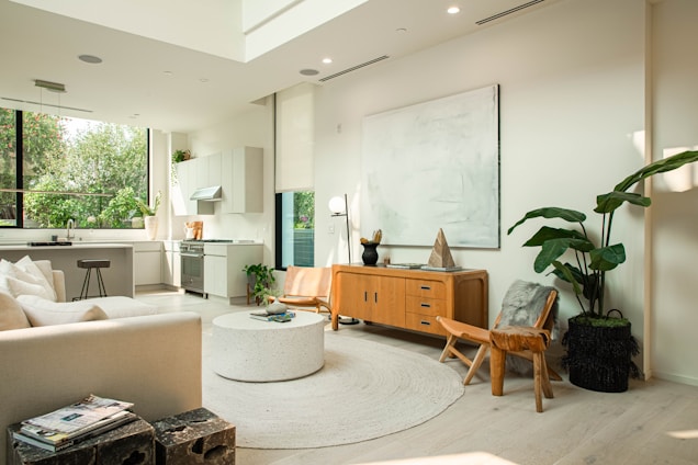 A modern, spacious living room with an open-plan kitchen. It features a light color palette with neutral tones. The kitchen area has sleek, minimalist cabinetry, a stove, and a large window allowing natural light to pour in. The living area includes a comfortable sofa, a circular coffee table, and a wooden sideboard with decorative items. Large leafy plants add a touch of greenery. The room is well-lit with a high ceiling and contemporary decor.