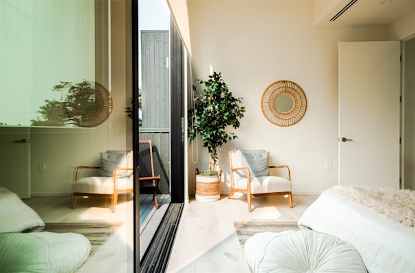 A bright, minimalist room with a cozy armchair and a decorative mirror on the wall. Potted plant adds greenery near a large glass door reflecting outdoors. The space includes a comfortable bed with fluffy pillows and a soft, textured throw. Light wooden flooring complements the neutral, calming color palette.