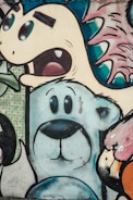 A vibrant mural featuring a variety of animated, cartoon-like characters. In the center, a blue and white bear with large, expressive eyes stands out. Above it, a dinosaur-like creature with spiky features and a surprised expression appears. To the right, part of a bird with a pink-orange gradient is visible. The artwork uses bold outlines and features a colorful mix of soft and bright hues.