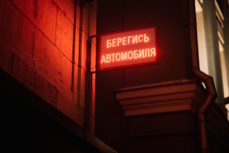 An illuminated red sign with Russian text is mounted on a corner of a building's exterior, casting a warm red glow on the stone wall. The urban setting is dimly lit, suggesting a night scene.
