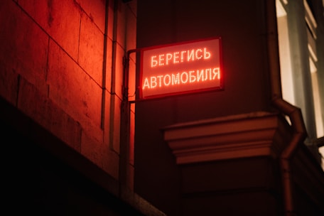 An illuminated red sign with Russian text is mounted on a corner of a building's exterior, casting a warm red glow on the stone wall. The urban setting is dimly lit, suggesting a night scene.