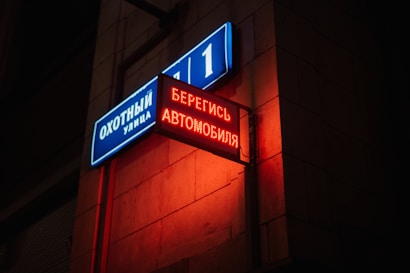 A street corner features two signs attached to a building. One sign is blue and white, labeled with street names and numbers in Cyrillic script. The other sign is a red, illuminated electronic message also in Cyrillic script, casting a vibrant glow against the brick wall.