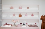 A minimalist store display featuring a variety of handbags in soft shades of pink, brown, and beige arranged on white shelves. The lower part of the image shows a wooden counter with folded clothing, a vase with pink flowers, a framed picture, and shoes. To the right, a mannequin dressed in a brown button-up dress is partially visible.