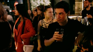 A group of people gather at a dimly lit bar, with one man prominently holding a drink and glancing to the side. Diverse individuals around him engage in conversation and enjoy the setting. The atmosphere is social and lively.