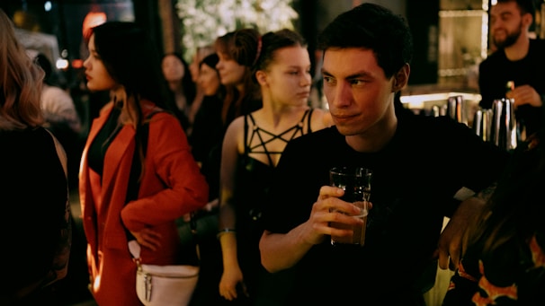 A group of people gather at a dimly lit bar, with one man prominently holding a drink and glancing to the side. Diverse individuals around him engage in conversation and enjoy the setting. The atmosphere is social and lively.