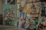 A collection of paintings displayed against a wall, each showcasing different subjects. The largest painting features vibrant flowers in red, yellow, and blue on a green background. Other paintings include koi fish amidst lily pads, a detailed village scene, and abstract art with various colors. Some of the paintings have a soft, naturalistic style, while others are more abstract and scenic.
