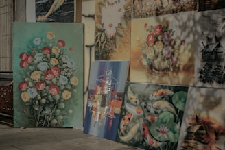 A collection of paintings displayed against a wall, each showcasing different subjects. The largest painting features vibrant flowers in red, yellow, and blue on a green background. Other paintings include koi fish amidst lily pads, a detailed village scene, and abstract art with various colors. Some of the paintings have a soft, naturalistic style, while others are more abstract and scenic.