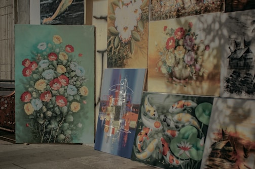 A collection of paintings displayed against a wall, each showcasing different subjects. The largest painting features vibrant flowers in red, yellow, and blue on a green background. Other paintings include koi fish amidst lily pads, a detailed village scene, and abstract art with various colors. Some of the paintings have a soft, naturalistic style, while others are more abstract and scenic.