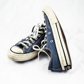 A pair of well-worn blue canvas sneakers with white soles and laces, positioned against a plain background. The shoes show visible signs of wear, adding a vintage or casual feel.