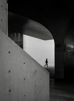 A solitary person is walking in a modern architectural space characterized by large concrete structures and a play of light and shadows. The area has a minimalist design with strong geometric shapes, creating a sense of depth and perspective.