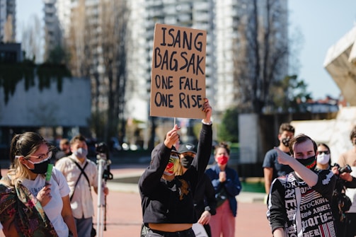 A group of people wearing face masks gather outdoors in an urban setting. One person holds a sign that reads 'Isang Bagsak One Fall One Rise.' The scene suggests a protest or rally, with people of diverse ages and backgrounds showing solidarity.