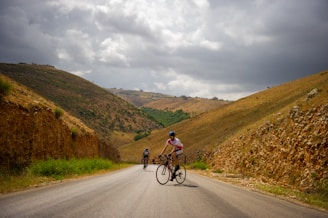 Two cyclists riding on a scenic country road with rolling hills under a bright sky.