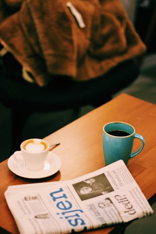 A cozy setting on a wooden table featuring a cappuccino in a white cup and saucer with a spoon, a black coffee in a blue mug, and a folded newspaper. A soft, brown blanket or jacket rests on a nearby chair, adding warmth to the scene.