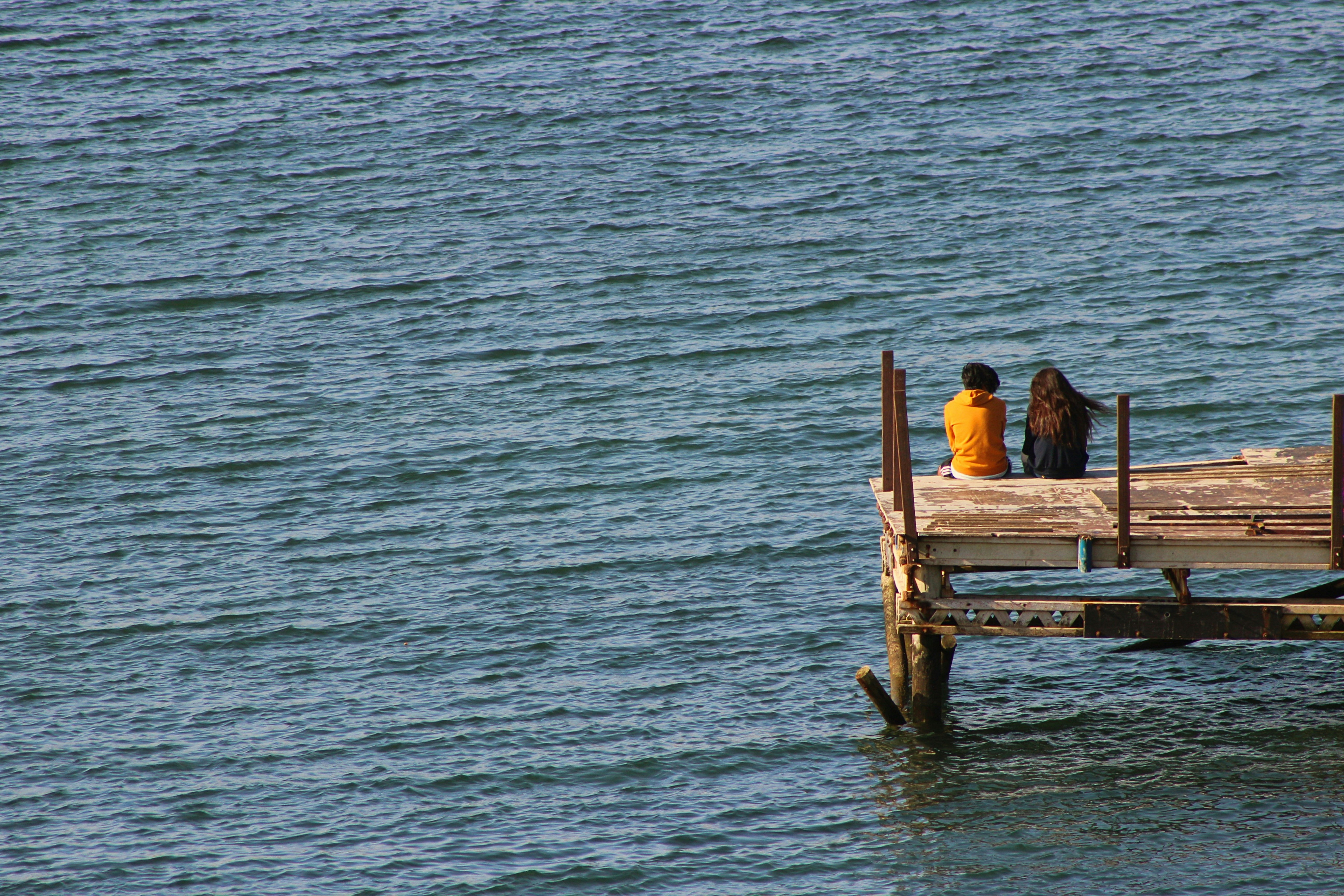 2 person sitting on brown wooden bench on sea water during daytime