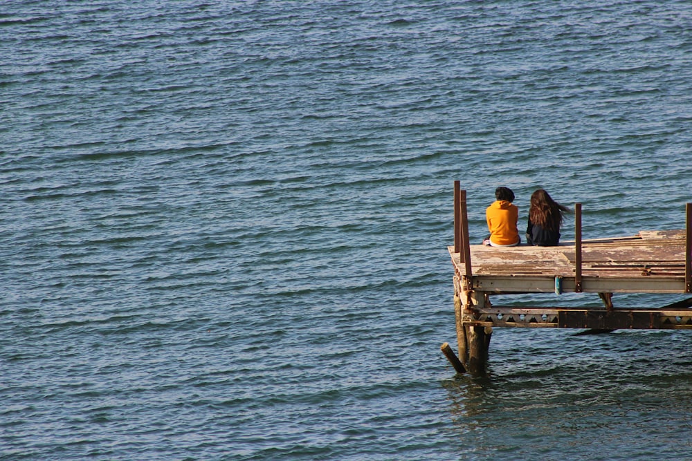 2 personnes assises sur un banc en bois brun sur l’eau de mer pendant la journée