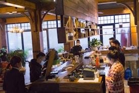 A cozy café interior features several people gathered around a wooden bar. Shelves above the bar are stocked with jars and other items. Large windows in the background allow natural light to illuminate the space, and plants and wooden elements add warmth to the setting. People are engaged and appear to be either ordering or preparing drinks.