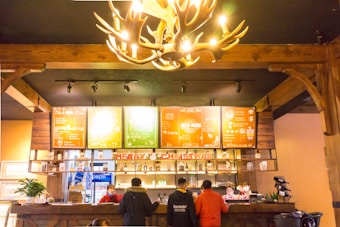 A cafe interior with warm wooden decor and a glowing antler chandelier overhead. The counter has Christmas decorations, including the words 'Merry Christmas'. Behind the counter are illuminated menu boards with a rustic design, and various beverages and items are neatly arranged on shelves. Several people are gathered at the counter, with staff in red uniforms attending to customers.