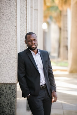 photography poses for men,how to photograph fortune vieyra wearing black business suit while standing next to a wall.; man in black suit jacket leaning on gray concrete wall during daytime