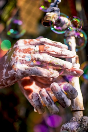 Hands covered in soap lather are positioned under a running faucet with foam bubbles floating around. The lighting creates a warm, colorful ambiance with hints of sunlight refracting through the bubbles.