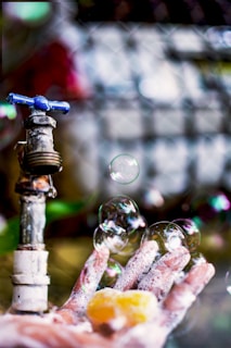 A close-up of a hand covered in soap foam is positioned under a weathered outdoor faucet. Several translucent bubbles float delicately above the hand, reflecting light and colors. The background is slightly blurred, enhancing the focus on the hand, faucet, and bubbles.