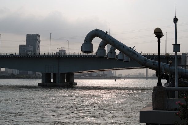An industrial bridge with large pipes extends over a river, reflecting light from the overcast sky. City buildings and power lines are visible in the background, and there are street lamps in the foreground.