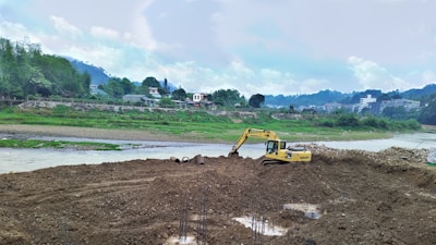 An excavator is working on a mound of soil near a riverbank. The background includes a village with various residential buildings and lush greenery on rolling hills. The sky is overcast with clouds.