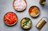 Several bowls are arranged on a surface, each filled with different cooking ingredients. There are chopped onions, diced tomatoes, an assortment of ground spices, chopped green chilies with garlic, and a bottle of oil. A grater is positioned nearby, completing the cooking setup.