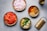 Several bowls are arranged on a surface, each filled with different cooking ingredients. There are chopped onions, diced tomatoes, an assortment of ground spices, chopped green chilies with garlic, and a bottle of oil. A grater is positioned nearby, completing the cooking setup.